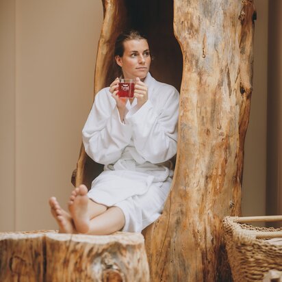 Entspannte Frau im Bademantel trinkt Tee in einem ausgehöhlten Baumstamm.